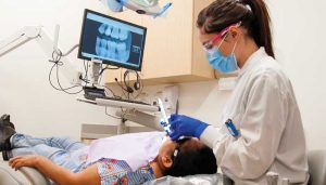 Dental provider cleans teeth of a patient reclining in dental chair, below TV monitor showing an xray of teeth.
